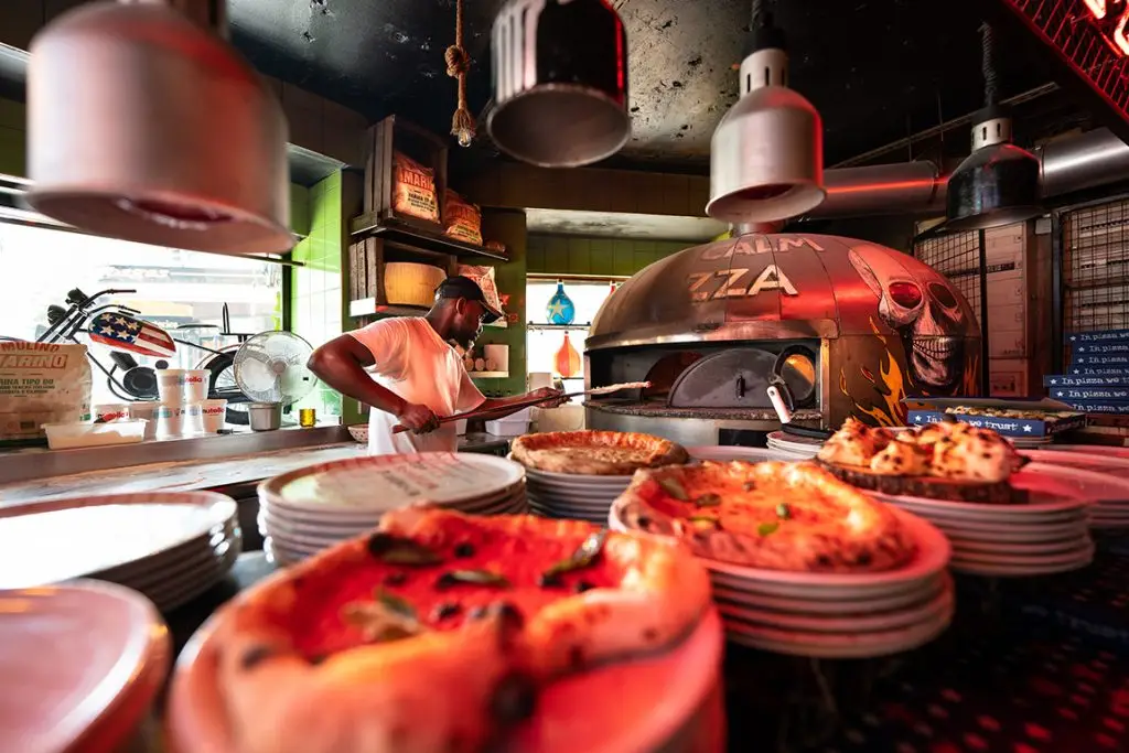 Photo d'une personne mettant une pizza dans un four a pizza. Au premier plan il y a des pizza sur des assiettes et au segond la personne. La personne est dans la cuisine d'un restaurant.