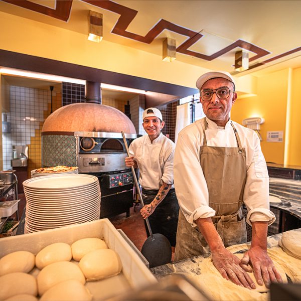 Photo de deux personne travaillant dans la cuisine d'une pizzeria. Une personne a a la main un outil qui permet de mettre la pizza dans le four et l'autre travaille la pate a pizza.