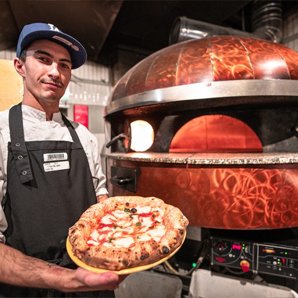 Photo d'une personne souriante avec une pizza a la main avec en arrière plan un four à pizza